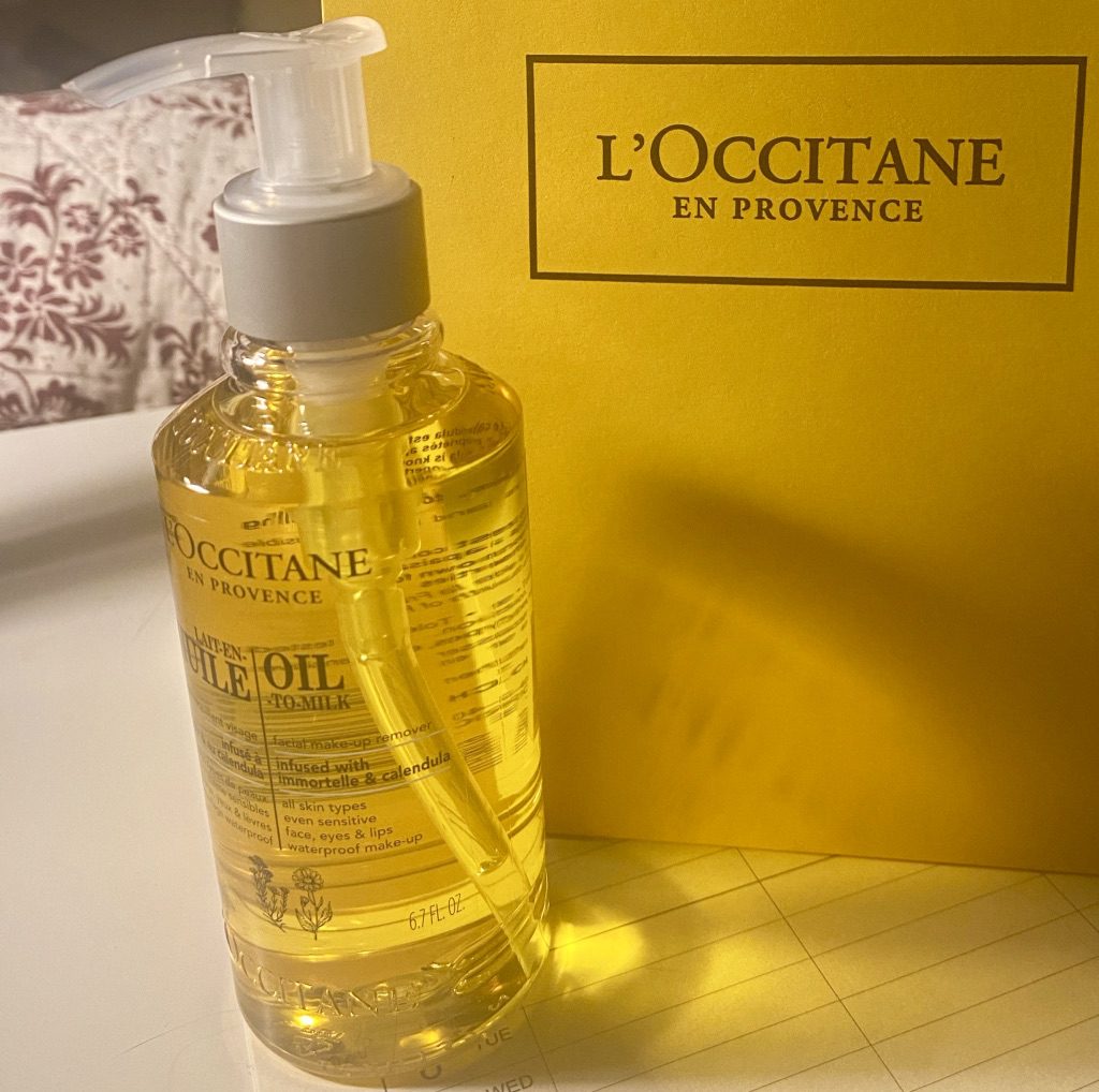 A full bottle of L'Occitane en Provence Oil to Milk Facial make-up remover stands on a desk next to the bag it was brought home in. Both the bag and the contents of the bottle are a bright, golden yellow colour. Light hitting the bottle casts shadows on the bag.