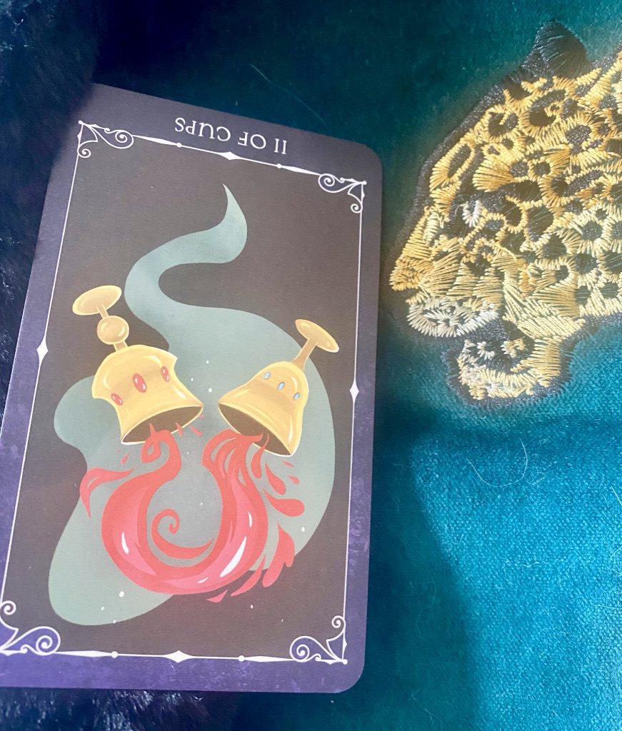 Embroidered leopard on green velvet looks incredulously at Disney Villains Tarot card showing two golden goblets overturned and spilling blood red wine